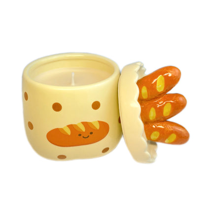 Baguette & butter scented candle in a ceramic jar with polka dots and a smiling baguette illustration, shown open with the baguette-shaped lid resting beside it on a white background.