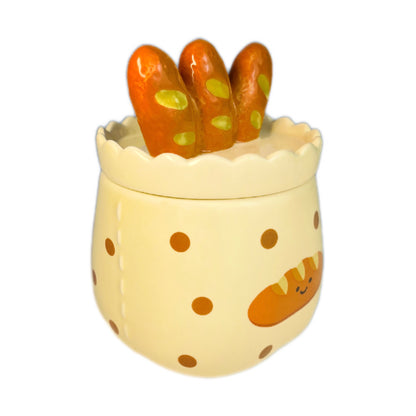 Baguette & butter scented candle in a ceramic jar with polka dots and a smiling baguette illustration, topped with a baguette-shaped lid, photographed on a white background.