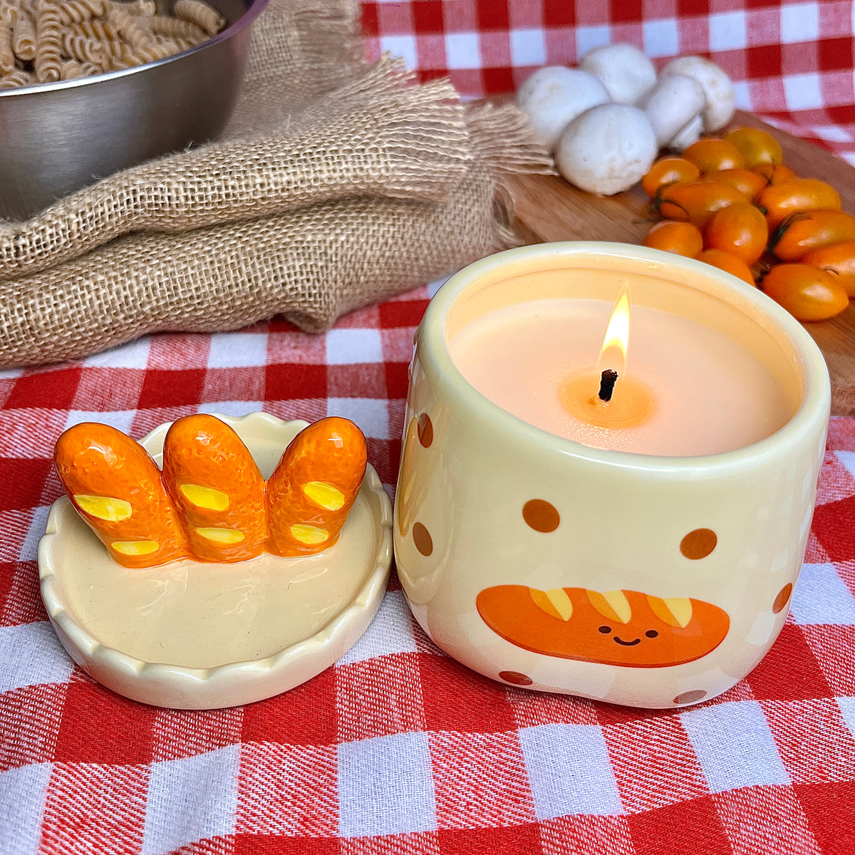 Lit baguette & butter scented candle in a ceramic jar with polka dots and a smiling baguette illustration, styled on a red gingham tablecloth with its baguette-shaped lid, fresh tomatoes, mushrooms, and kitchen linens in the background.
