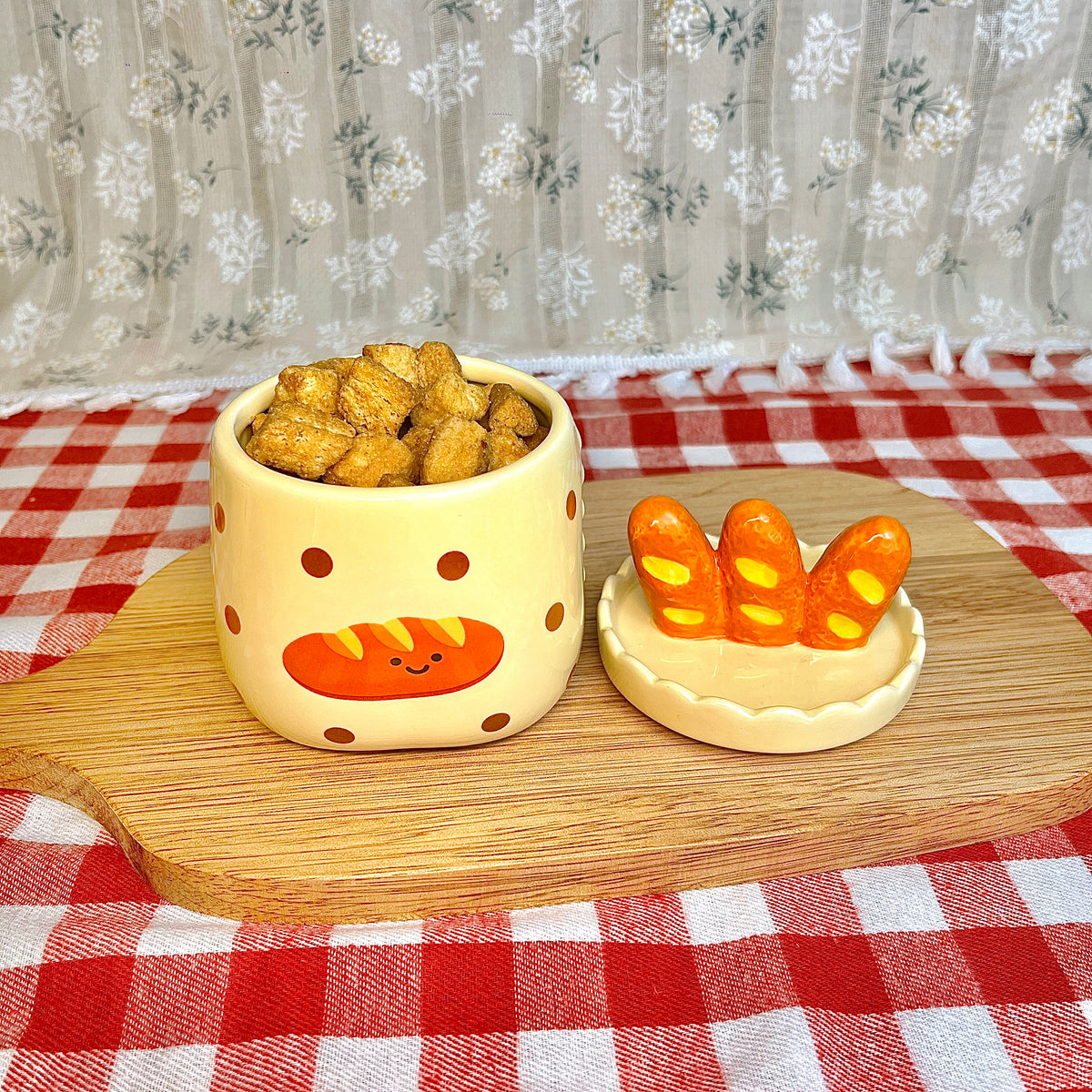 Repurposed ceramic candle jar with polka dots and a smiling baguette illustration, reused as a snack container filled with crackers, with the baguette-shaped lid placed beside it on a wooden board and red gingham tablecloth.