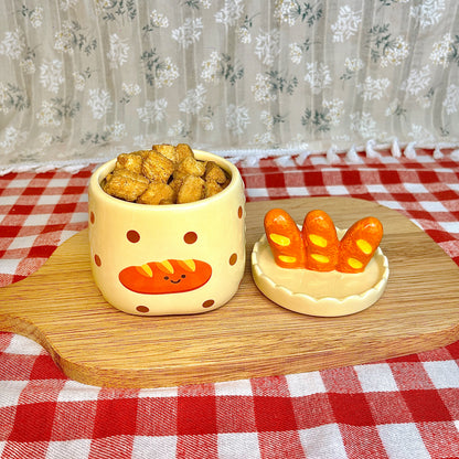 Repurposed ceramic candle jar with polka dots and a smiling baguette illustration, reused as a snack container filled with crackers, with the baguette-shaped lid placed beside it on a wooden board and red gingham tablecloth.