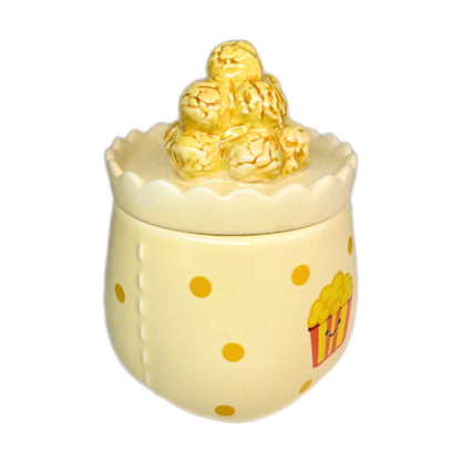 Caramel popcorn scented candle in a ceramic jar with yellow polka dots and a smiling popcorn bucket illustration, topped with a popcorn-shaped lid, photographed on a white background.