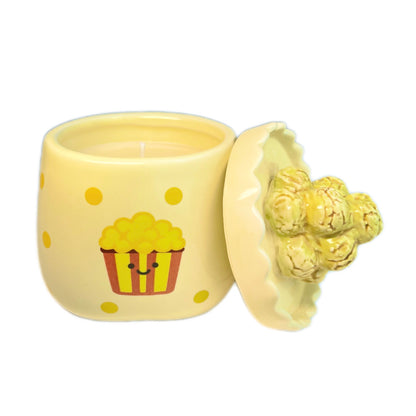 Caramel popcorn scented candle in a ceramic jar with yellow polka dots and a smiling popcorn bucket illustration, shown open with the popcorn-shaped lid resting beside it on a white background.