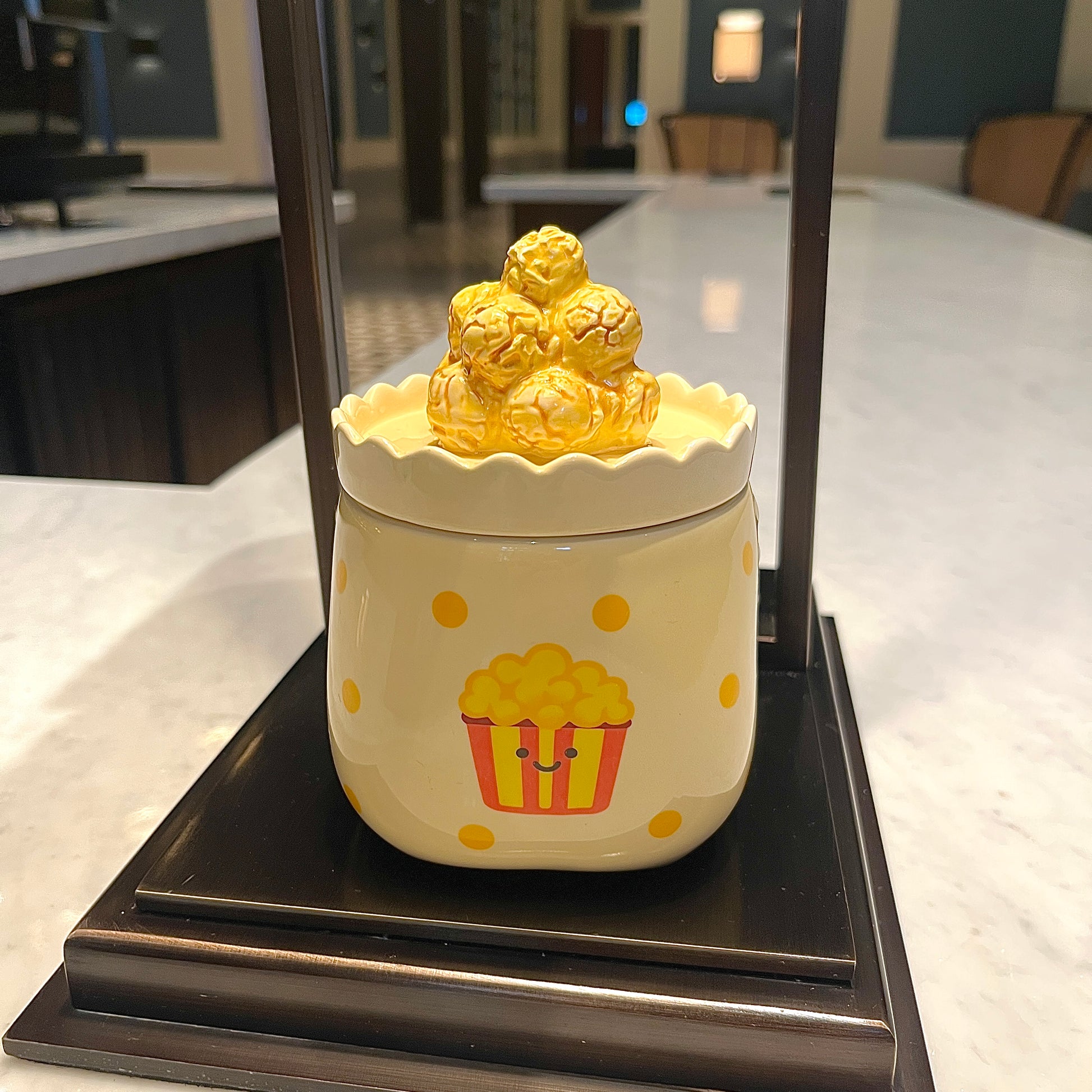 Caramel popcorn scented candle in a ceramic jar with yellow polka dots and a smiling popcorn bucket illustration, topped with a popcorn-shaped lid and displayed on a decorative stand in a modern kitchen setting.