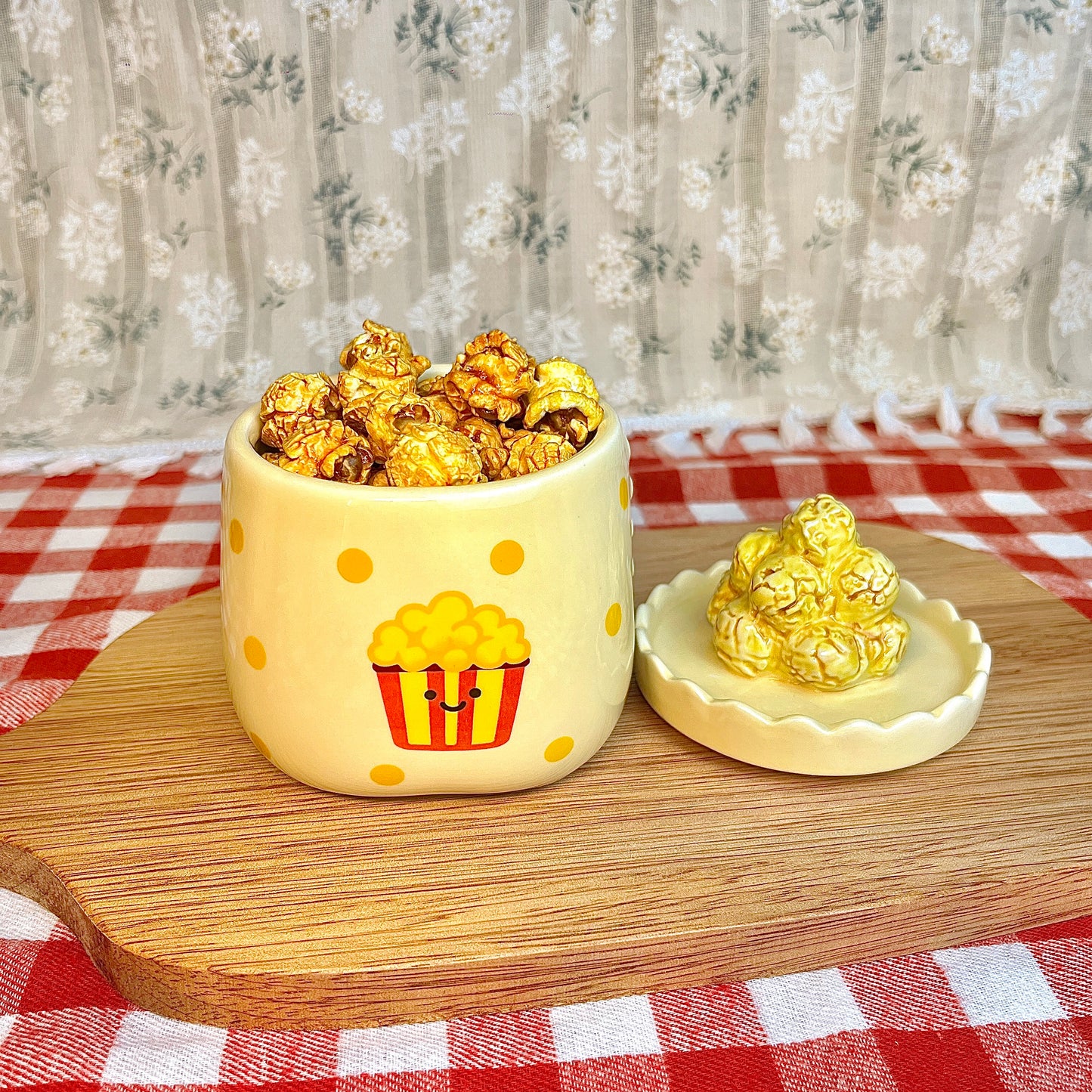 Repurposed ceramic candle jar with yellow polka dots and a smiling popcorn bucket illustration, reused as a snack cup filled with caramel popcorn, with the popcorn-shaped lid placed beside it on a wooden board and red gingham tablecloth.