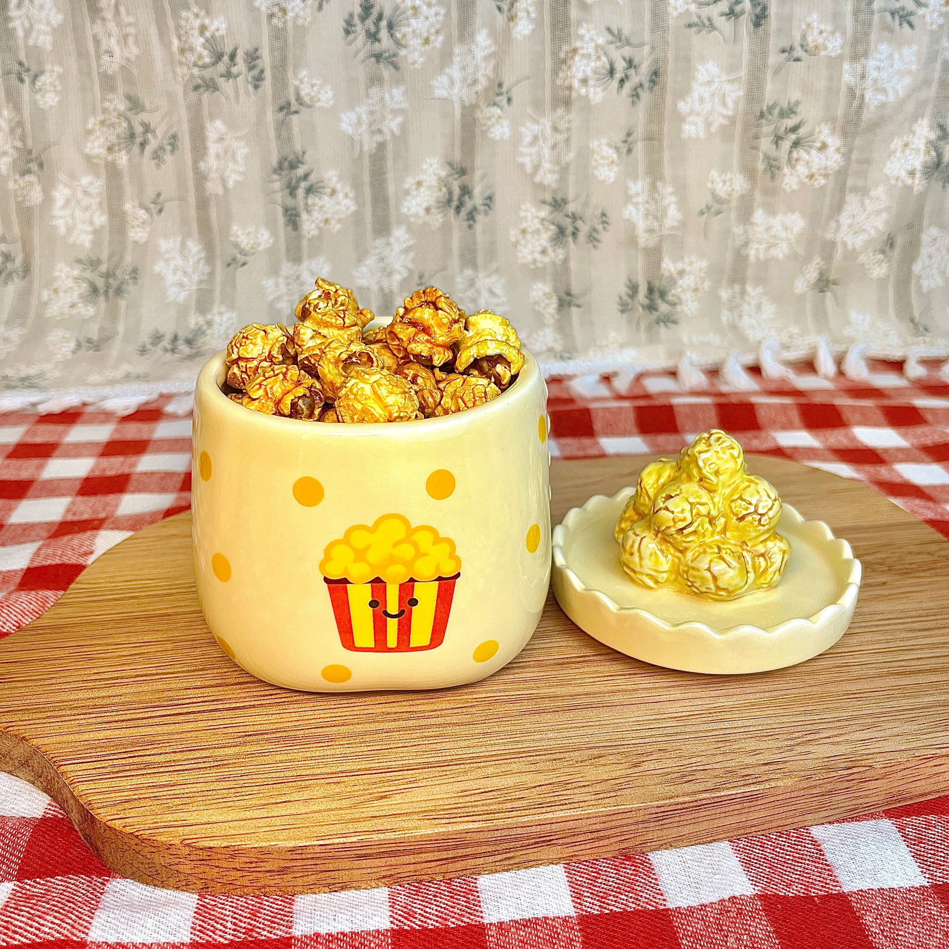 Repurposed ceramic candle jar with yellow polka dots and a smiling popcorn bucket illustration, reused as a snack cup filled with caramel popcorn, with the popcorn-shaped lid placed beside it on a wooden board and red gingham tablecloth.