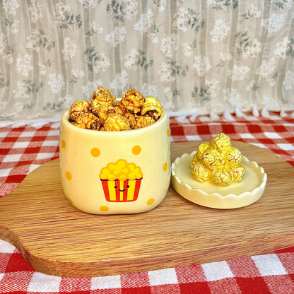 Repurposed ceramic candle jar with yellow polka dots and a smiling popcorn bucket illustration, reused as a snack cup filled with caramel popcorn, with the popcorn-shaped lid placed beside it on a wooden board and red gingham tablecloth.