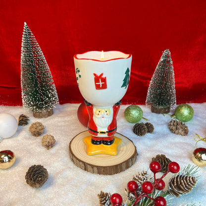 Spiced cranberry cider scented ceramic Santa goblet candle glowing in a festive Christmas scene with pinecones, ornaments, and snowy décor.