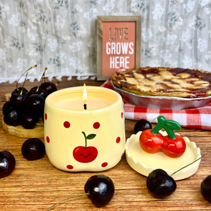 Lit velvet cherry scented candle in a ceramic jar with red polka dots and a smiling cherry illustration, styled on a wooden surface with its cherry-shaped lid, fresh cherries, and a cherry pie in the background.