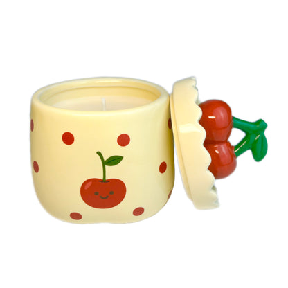Velvet cherry scented candle in a ceramic jar with red polka dots and a smiling cherry illustration, shown open with the cherry-shaped lid resting beside it on a white background.