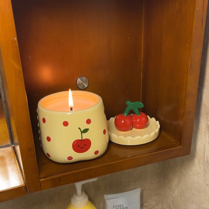 Lit velvet cherry scented candle in a ceramic jar with red polka dots and a smiling cherry illustration, displayed on a wooden shelf with its cherry-shaped lid placed beside it.