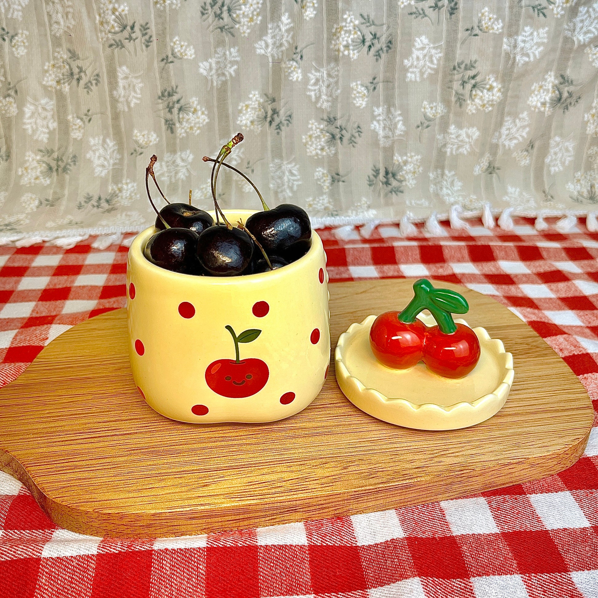 Repurposed ceramic candle jar with red polka dots and a smiling cherry illustration, reused as a small bowl holding fresh cherries, with the cherry-shaped lid placed beside it on a wooden board and red gingham tablecloth.