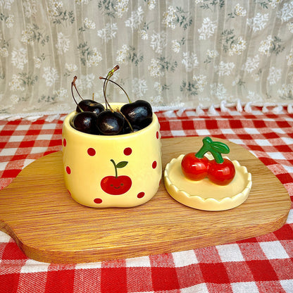 Repurposed ceramic candle jar with red polka dots and a smiling cherry illustration, reused as a small bowl holding fresh cherries, with the cherry-shaped lid placed beside it on a wooden board and red gingham tablecloth.